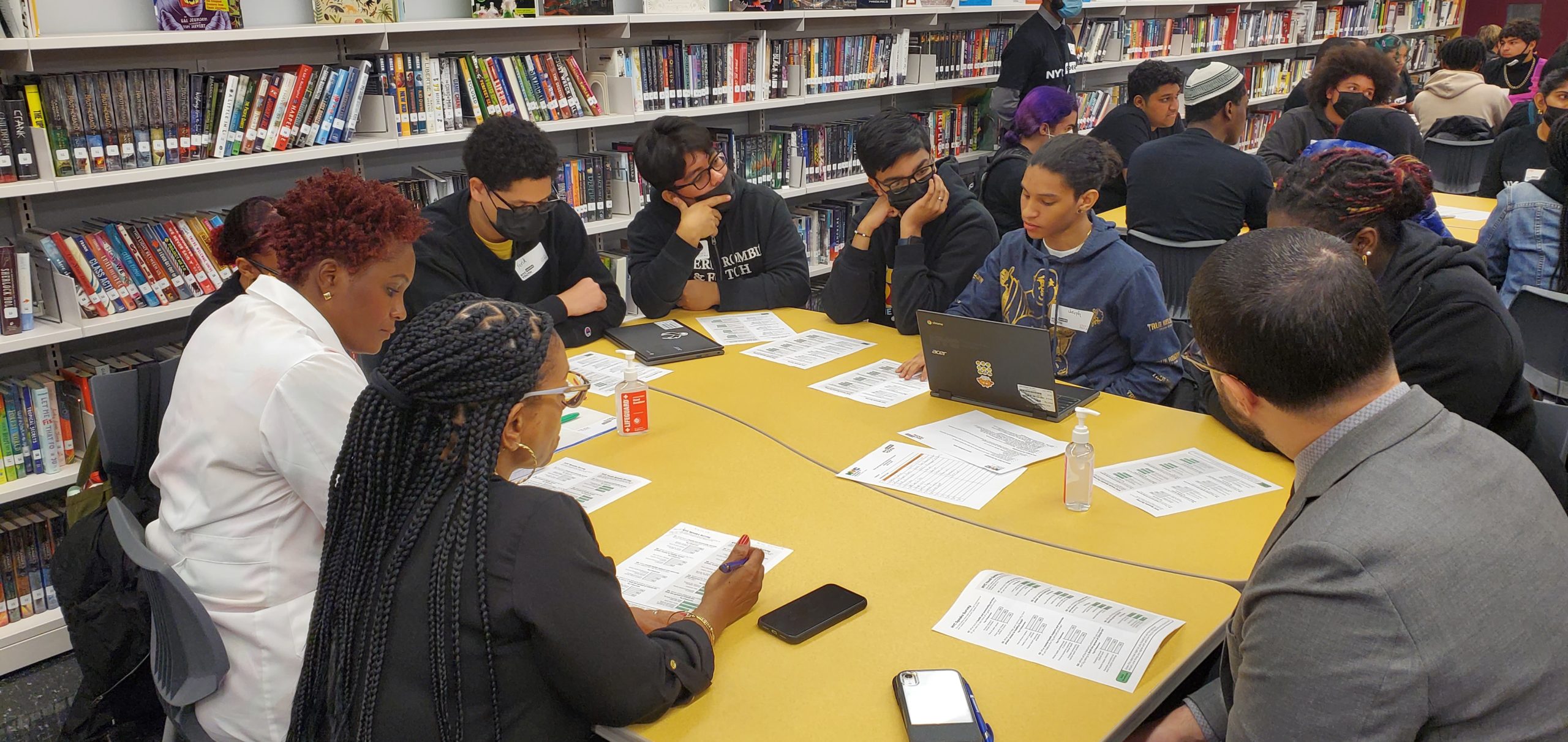 A group of Black and brown teenagers sits around two yellow tables pushed together, with one adult joining them. They all have NYC Speaks data handouts in front of them. One student has a computer open, facilitating the conversation. Behind them is a wall of bookshelves filled with books.