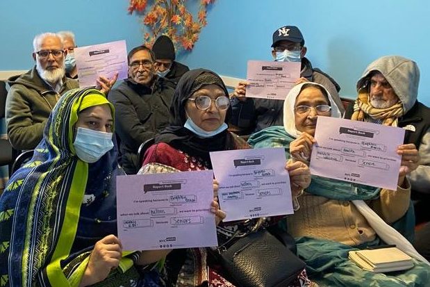 A group of older South Asian men and women, some wearing a hijab, hold up a NYC Speaks Community Conversation sheet showing their takeaways. They are seated close together in a bright blue room.